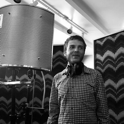 Man in plaid shirt in recording studio, smiling, headphones around neck.