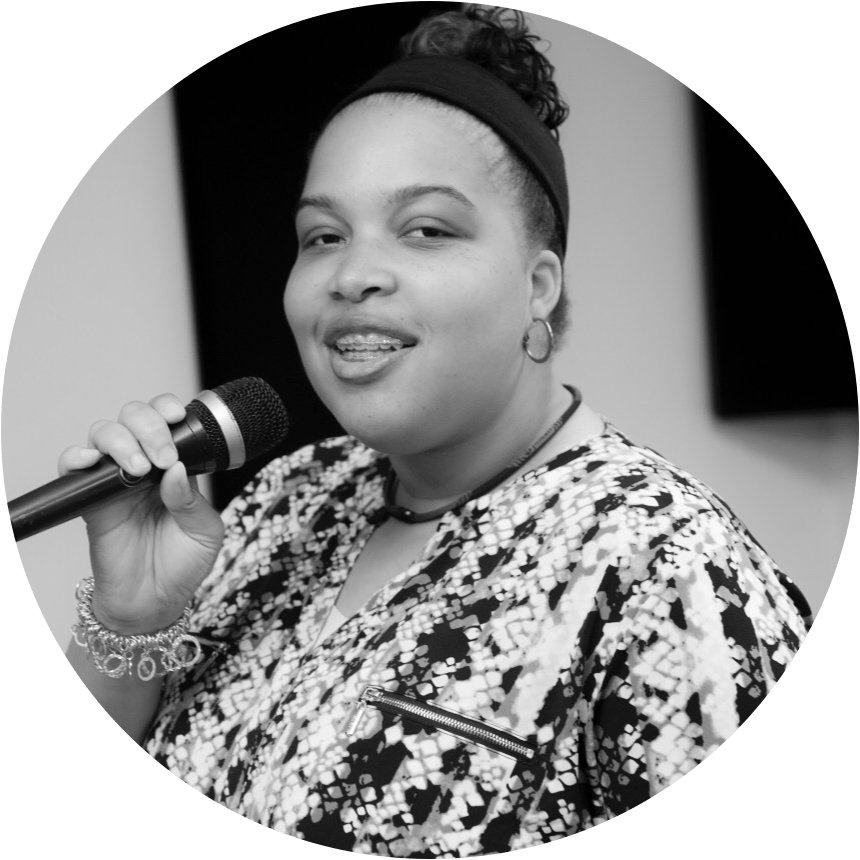 Woman with microphone, wearing a patterned shirt, black headband, and braces. Smiling, inside a room.