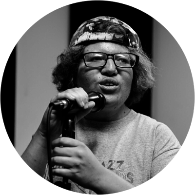 Person with glasses, hat, and microphone singing on stage. Black and white.