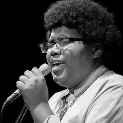 Black and white photo of a person with an afro singing into a microphone, wearing glasses and a collared shirt.