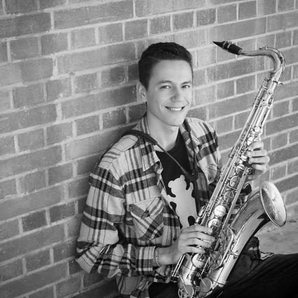 Smiling young man with saxophone, leaning against a brick wall. Wearing plaid shirt and black t-shirt.