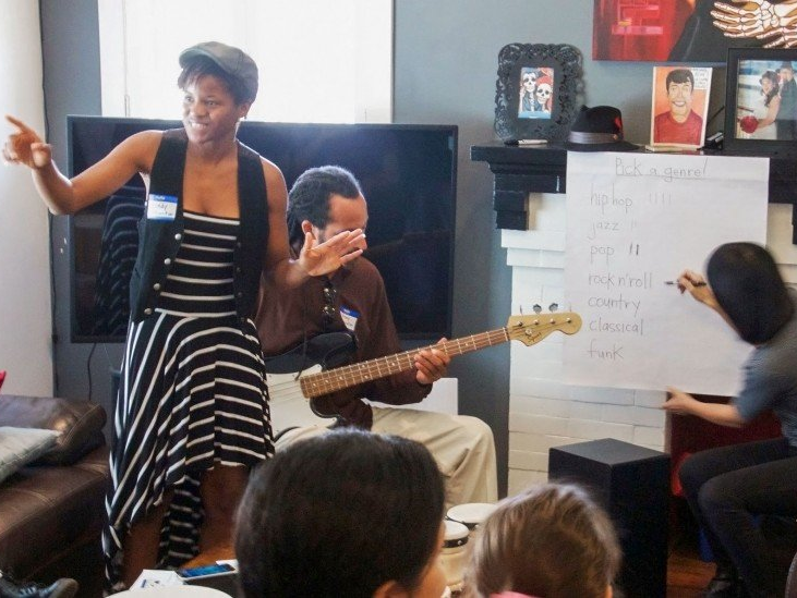 Woman gesturing, teaching; man with bass guitar; another writing on whiteboard; workshop setting.
