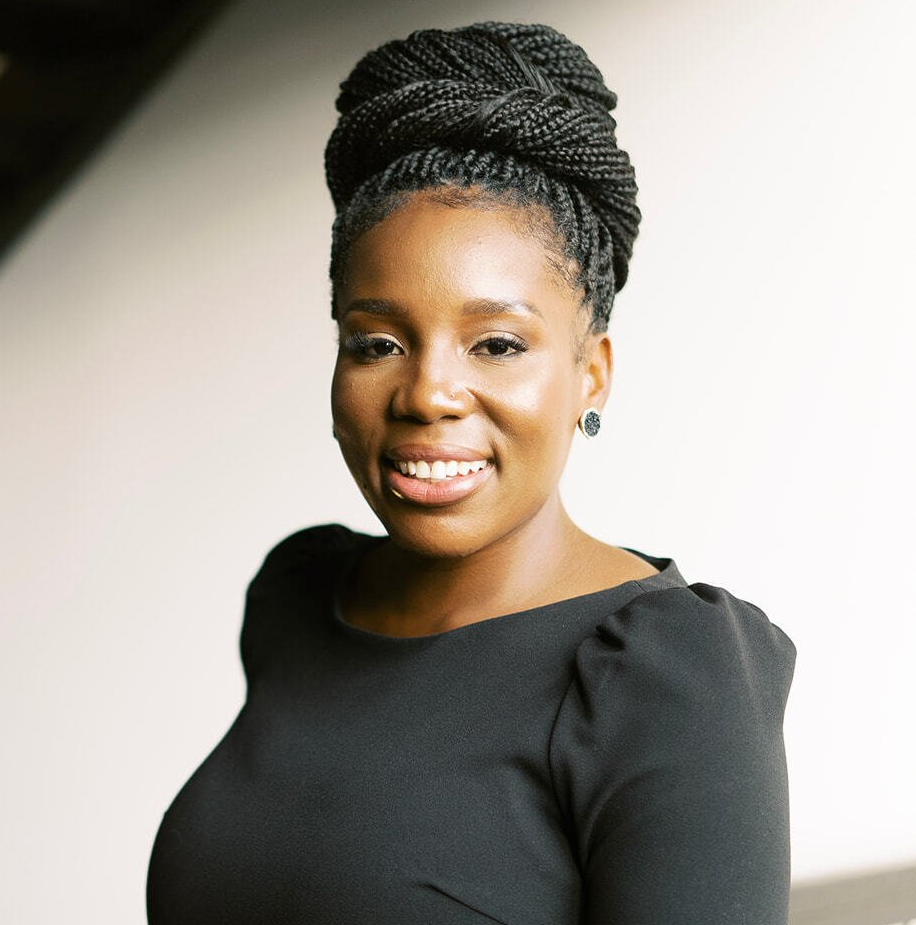 Black woman with braided updo smiles, wearing a black dress. Light background.