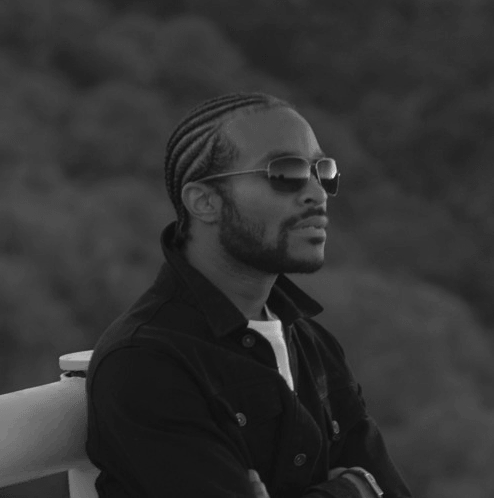 Man with braided hair and sunglasses, arms crossed, looking off to the side. Black and white photo.