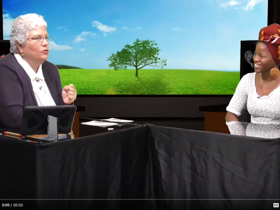 Two women in a studio, one speaking, one smiling, backdrop of a tree in a green field.