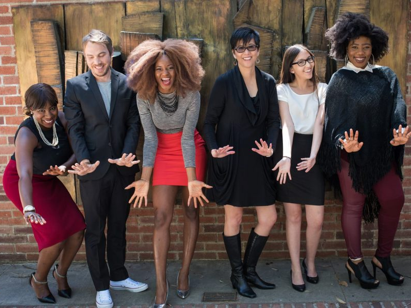 Six diverse people, arms outstretched, smiling in front of a brick wall.