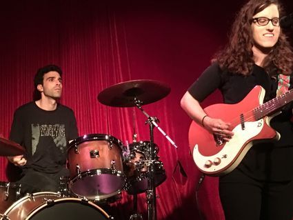 Drummer and guitarist on stage at a venue with red backdrop. The guitarist is playing and wearing glasses.