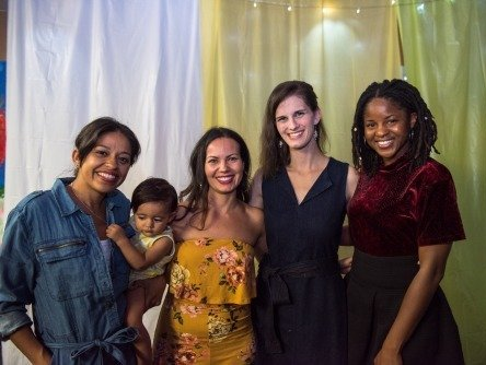 Five smiling people pose together, including a woman holding a baby. The background is decorated with fabric and lights.