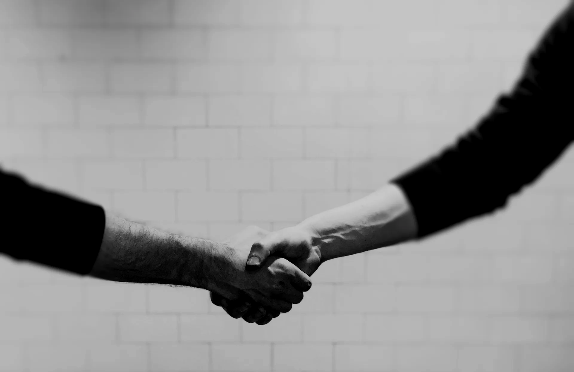 A black and white close-up of two people firmly shaking hands against a plain, tiled wall background.