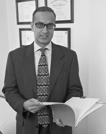 Dr. Manish Tandon Man in suit, glasses, holding a file; office setting, diplomas on the wall.