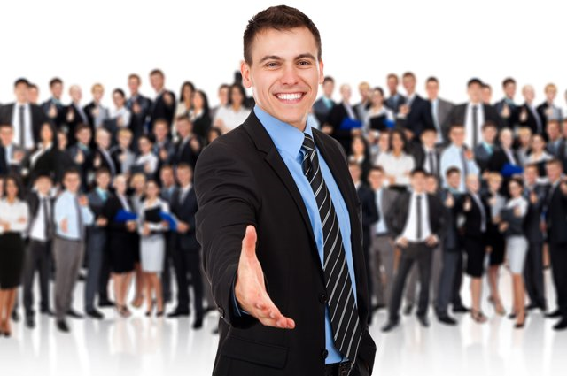 A business man in a suit offers the viewer a handshake while standing in front of a large group of his collegues