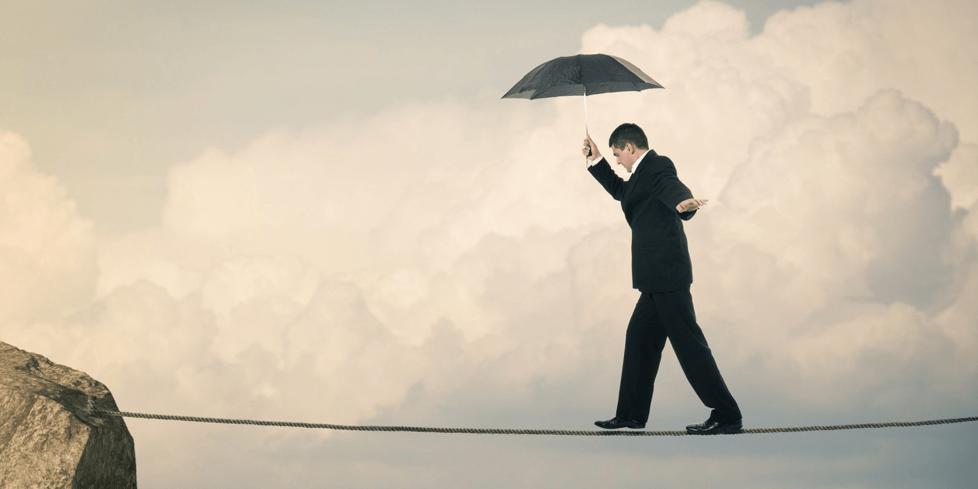 A man wearing a suit and holding an umbrella cautiously walks a tightrope.