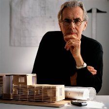 Man with glasses and dark shirt, seated at a table with architectural models and blueprints, contemplating, indoors.