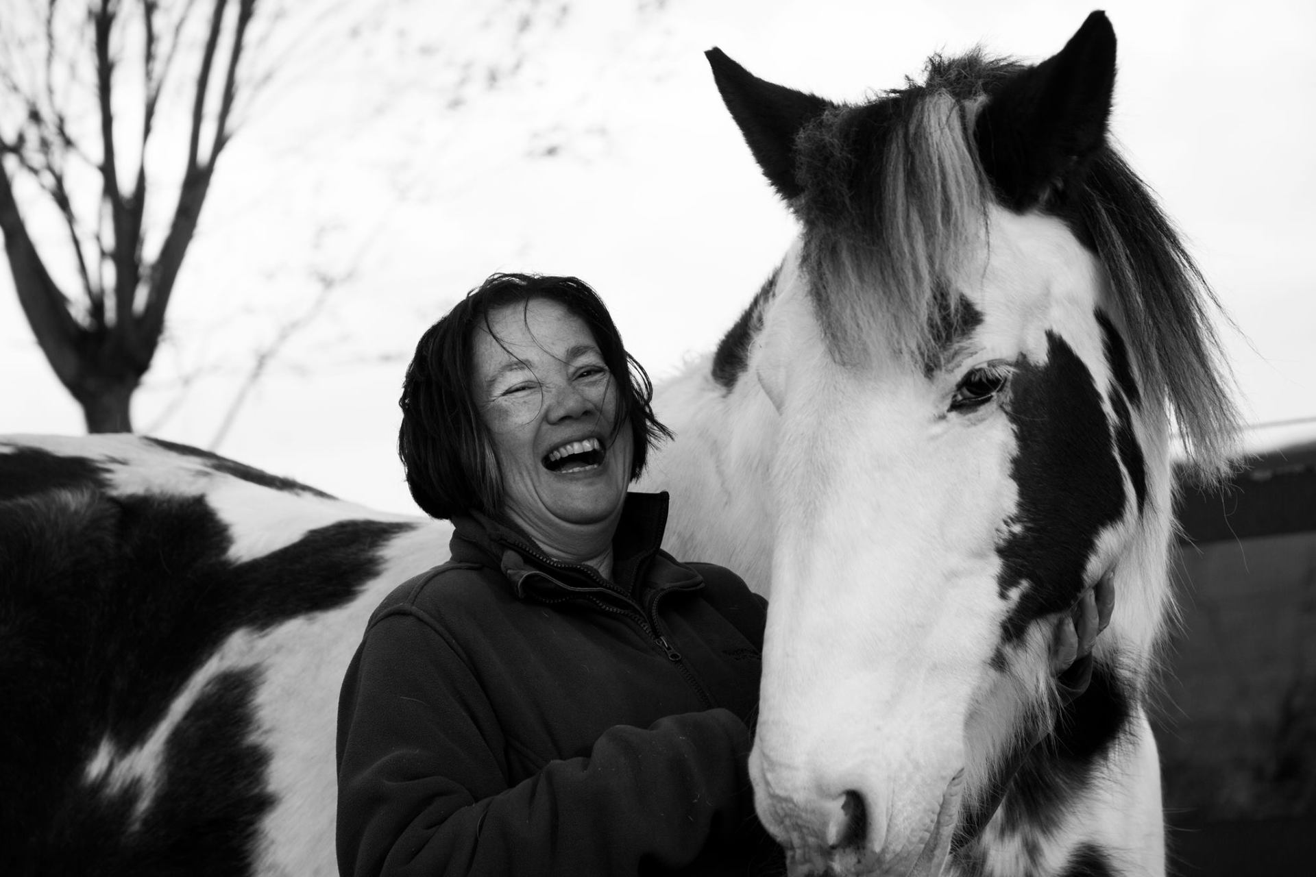 Een vrouw lacht naast een zwart-wit paard, buiten. Het paard heeft een gevlekte vacht.