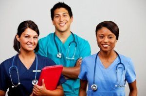Three smiling healthcare workers in scrubs standing together, one holding a red clipboard, with stethoscopes visible