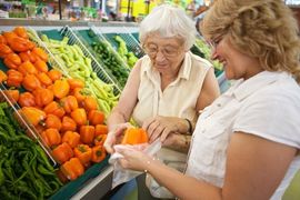 Two shoppers comparing produce in a grocery store, holding oranges near a display of vegetables.