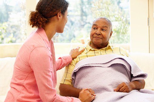 Caregiver comforting a patient in bed, holding her hand in a bright hospital room