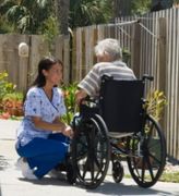 Nurse kneels beside a wheelchair user on a sunny garden walkway, speaking with them.