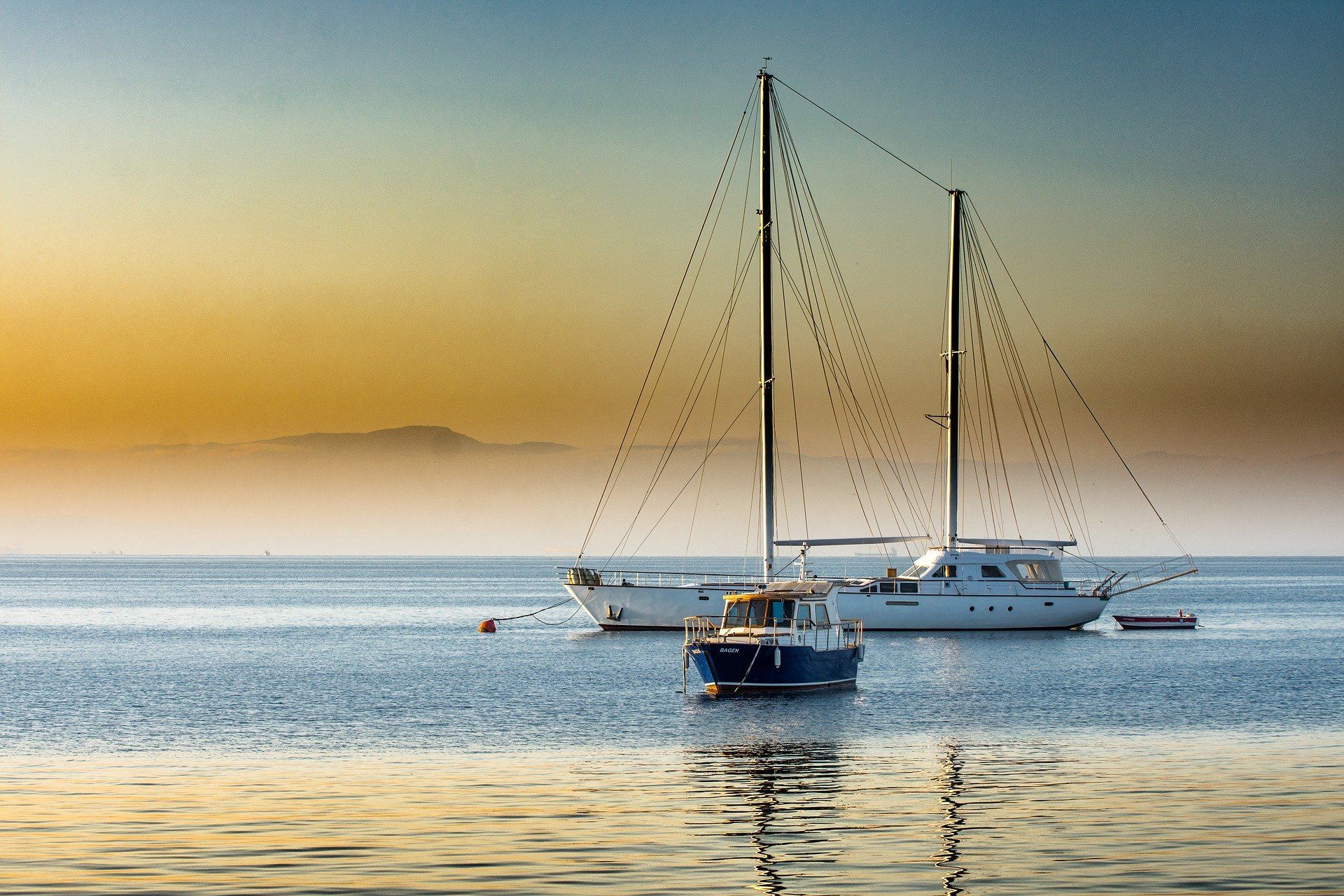 Sailboats on calm water at sunrise, distant island shrouded in mist, orange sky.