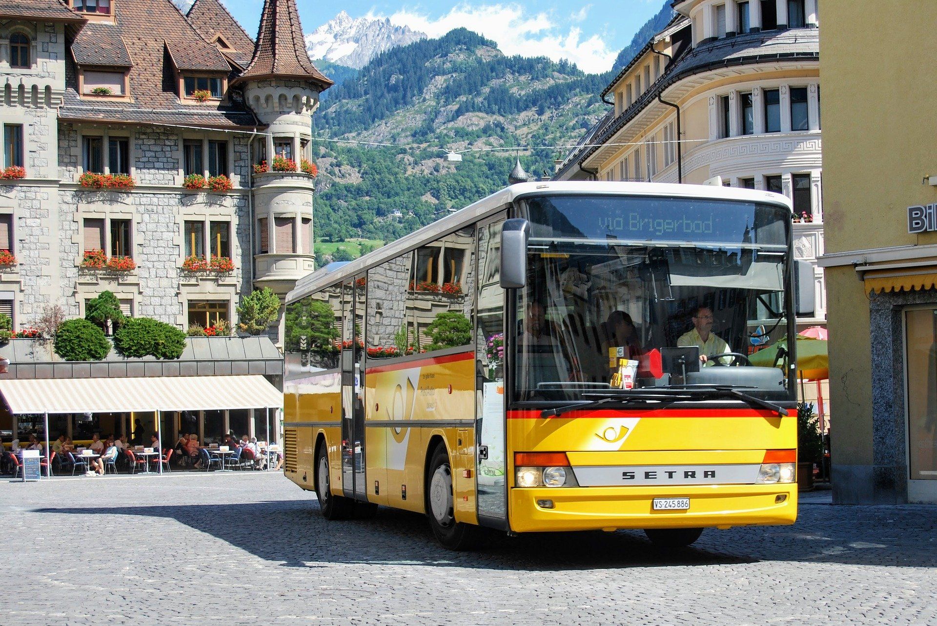 Yellow bus driving on cobblestone street in a town with buildings and mountain backdrop.