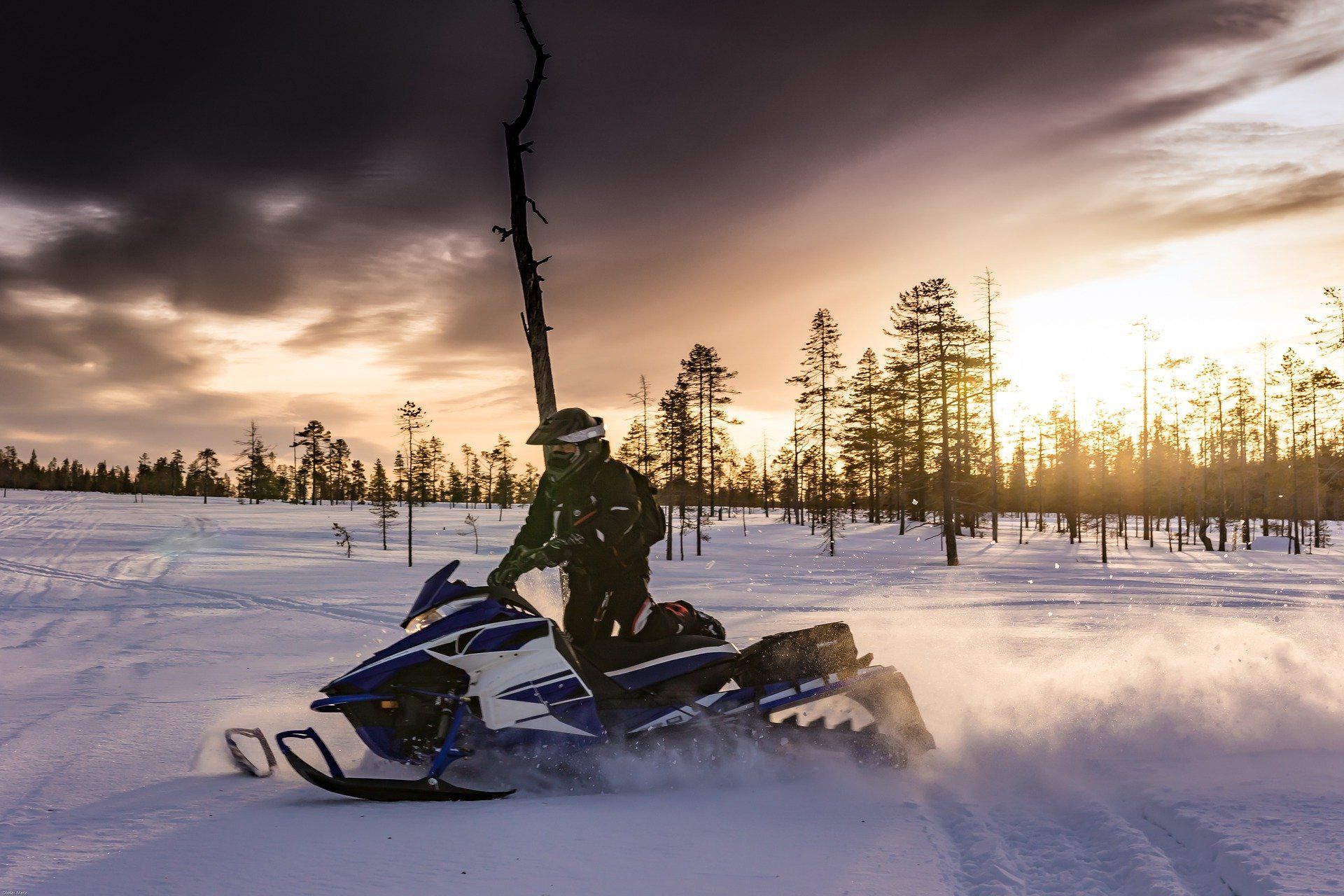 Snowmobiler on a blue and white snowmobile in a snowy landscape, kicking up snow, with a sunset in the background.