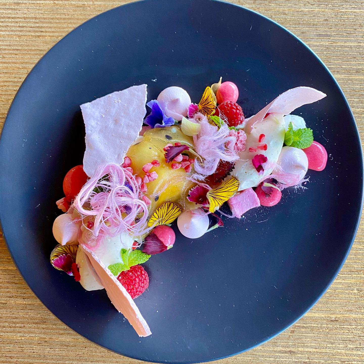 A black plate topped with fruit and flowers on a wooden table.