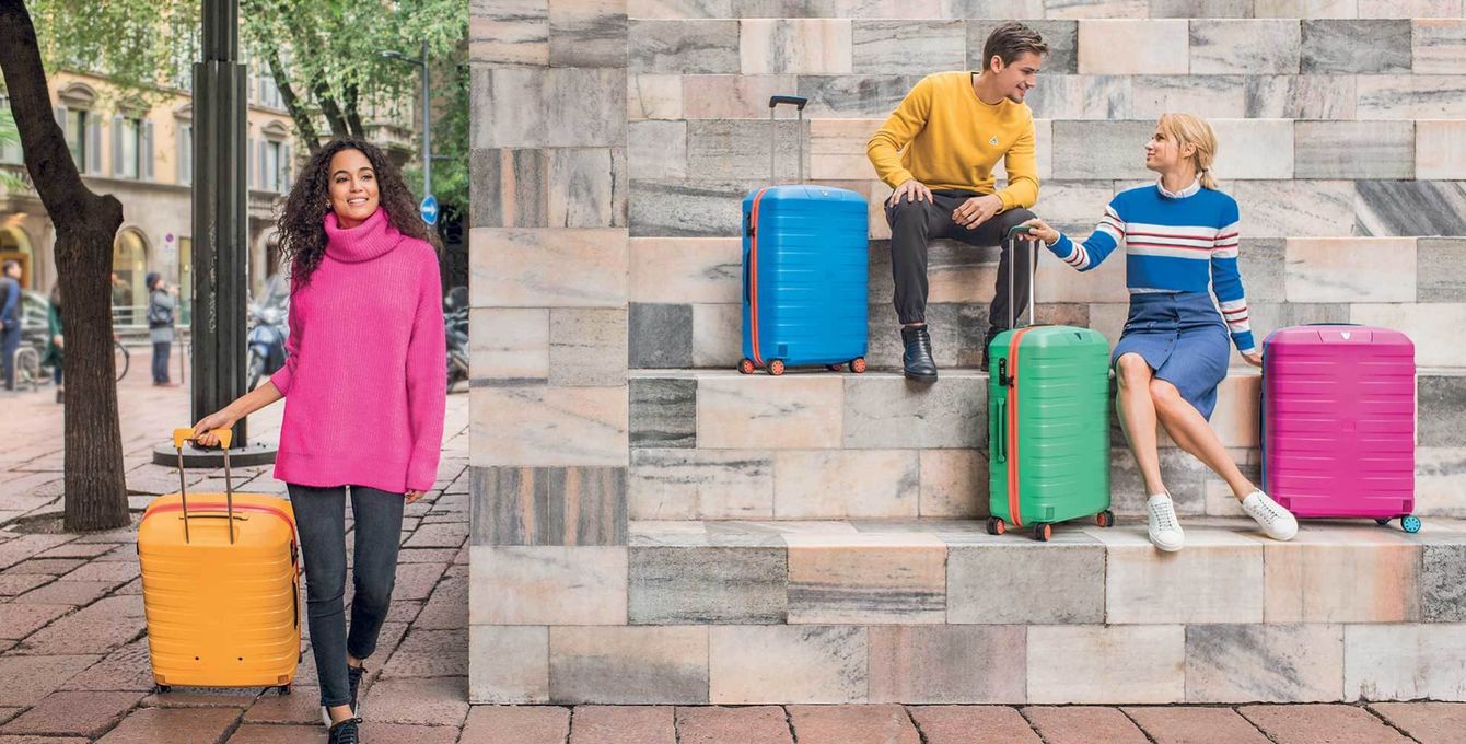 Viaggiatori con valigie colorate in piedi e seduti accanto a un muro di pietra in una strada cittadina