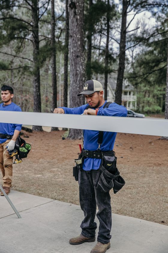 Two workers in blue long-sleeve shirts and tool belts lift a long, white wooden beam outdoors near a forest.
