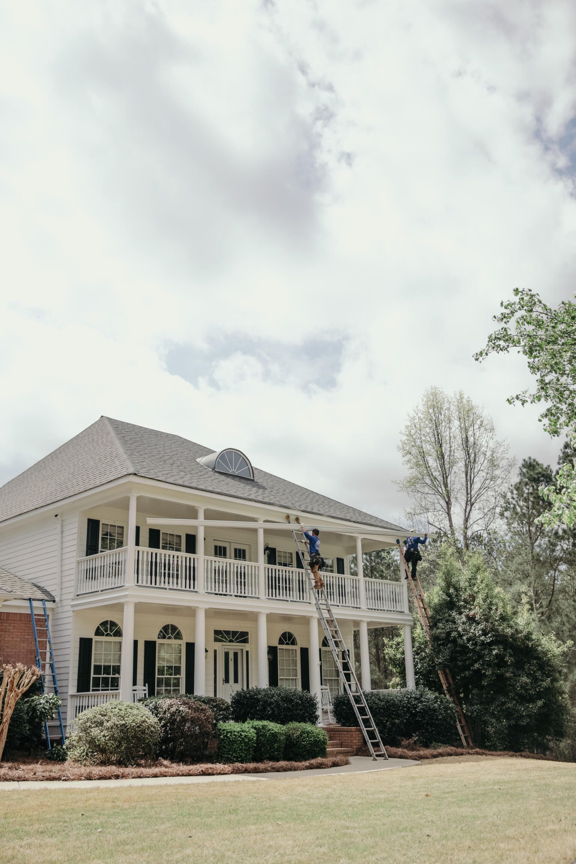Two people stand on ladders against a large, two-story white house with a porch and black shutters.