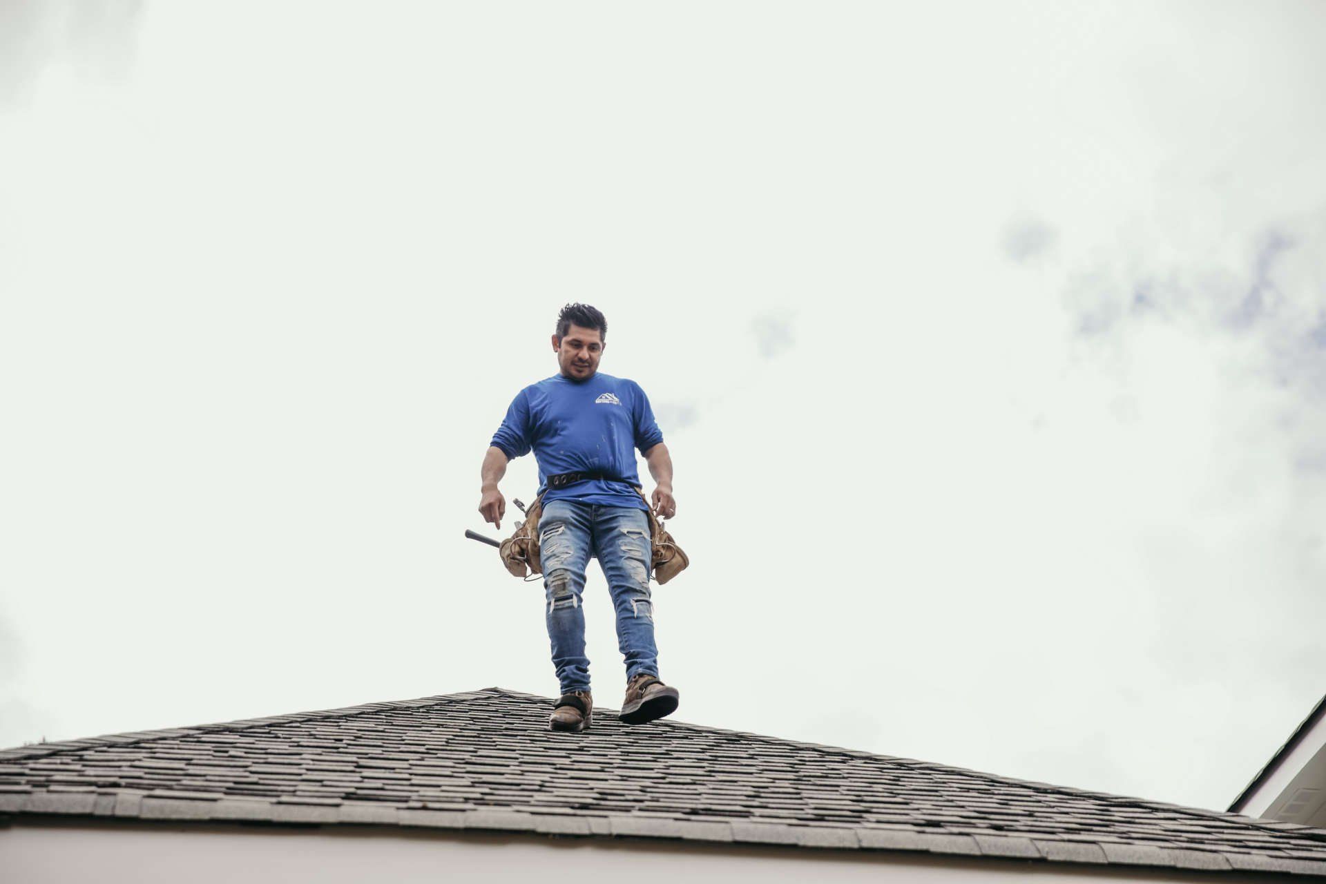 A person in a blue shirt and work jeans stands on a shingled roof, wearing a tool belt.