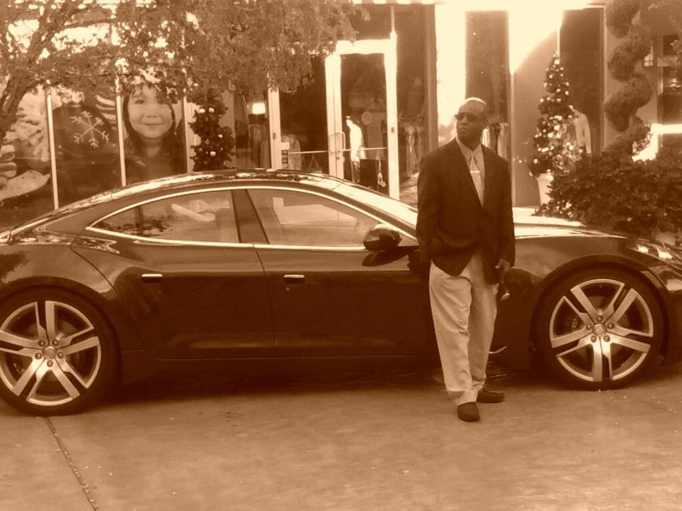 Man standing beside a black sports coupe parked in front of a building at sunset