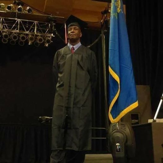 Graduate in cap and gown stands onstage beside a blue flag