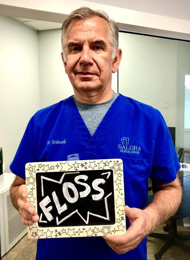 Man in blue scrubs holding a framed sign that says “FLOSS” in a dental office