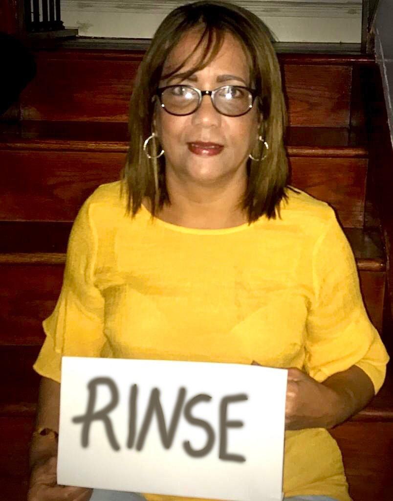 Person seated on wooden stairs holding a sign that reads “RIWSE,” wearing a yellow top and glasses.