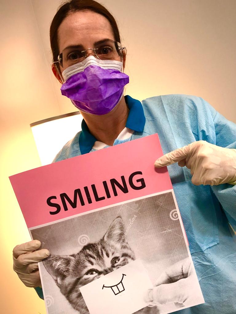 Masked person holding a pink sign that says “SMILING” above a cat drawing with a toothy grin