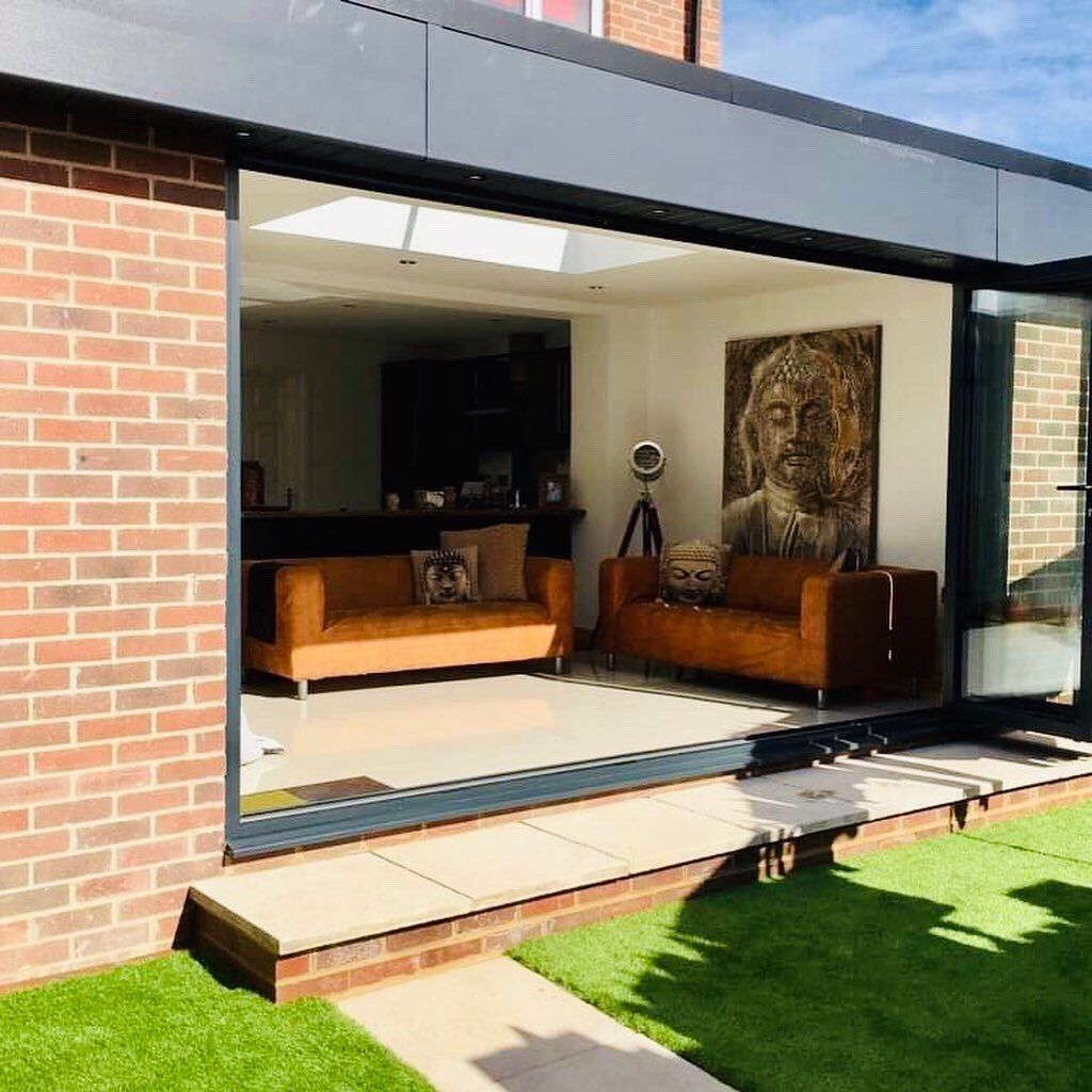 A modern home extension with open glass doors leading to a living room with tan sofas and a large Buddha wall portrait.