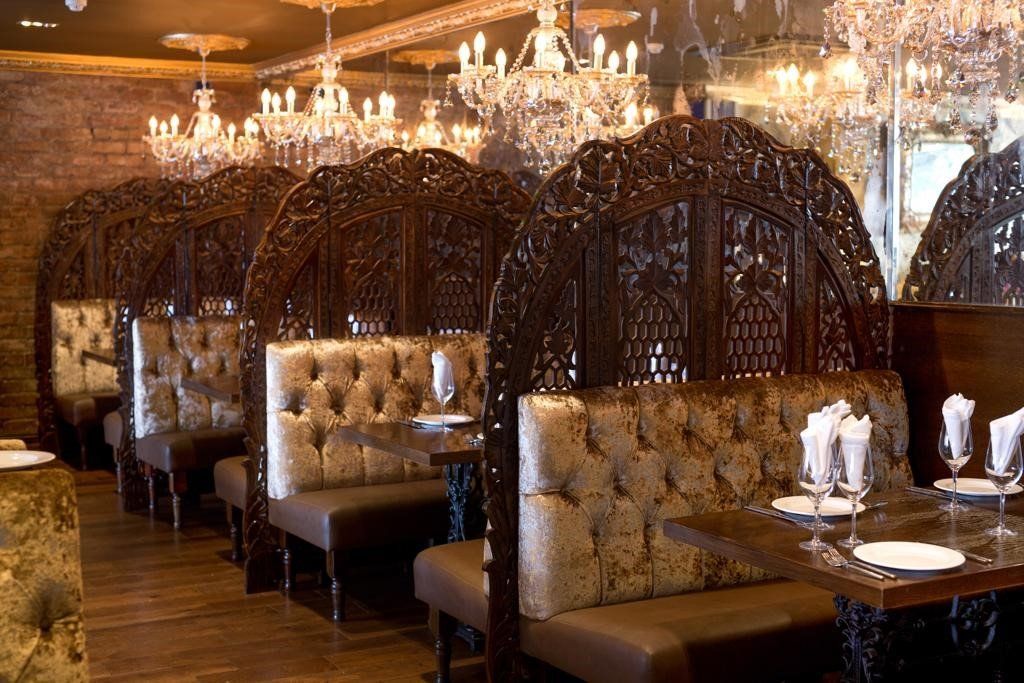 A dimly lit restaurant interior with plush, tufted booths separated by ornate, dark wood carved screens under chandeliers.