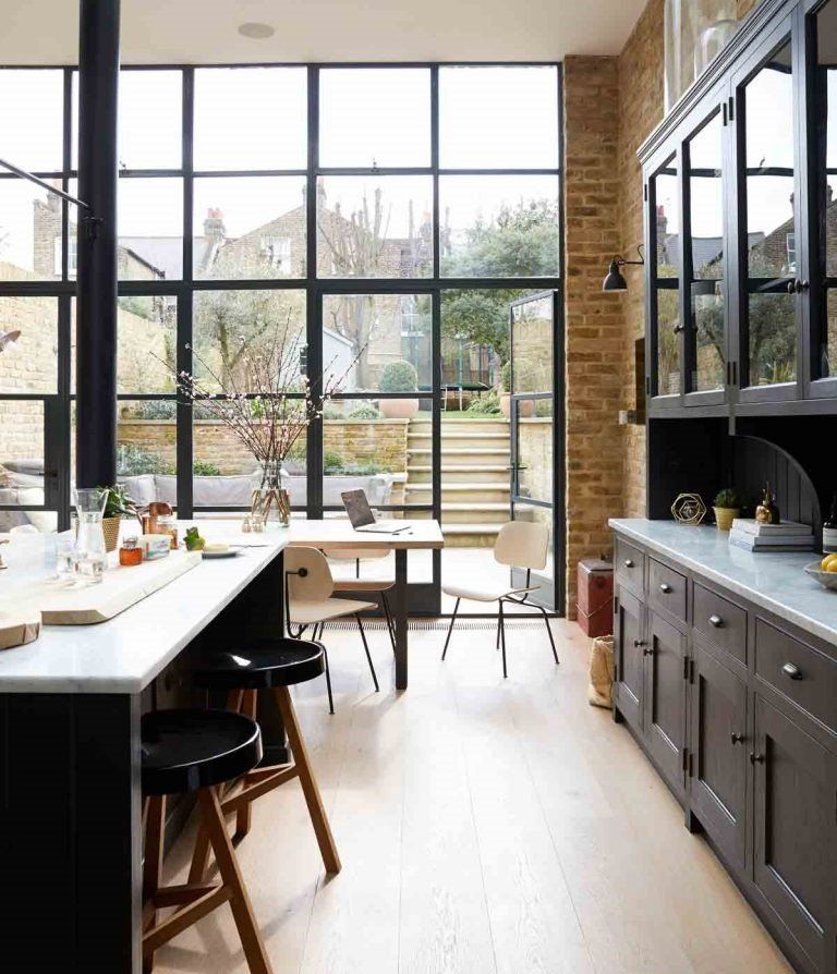 A modern kitchen with black cabinets, a white marble island, and a large floor-to-ceiling glass wall overlooking a garden.