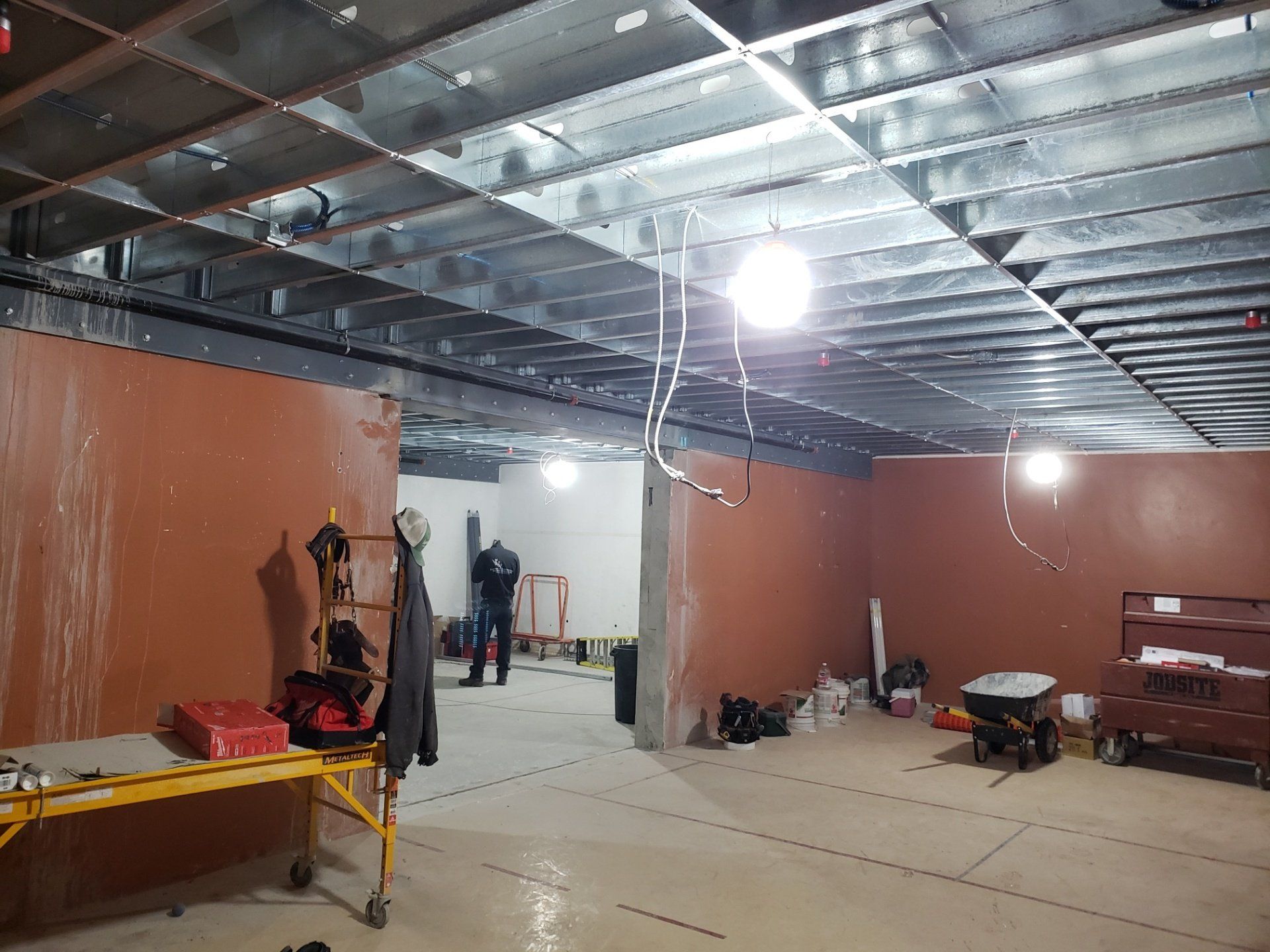 Unfinished indoor construction space with exposed metal ceiling, hanging lights, and scattered tools and materials