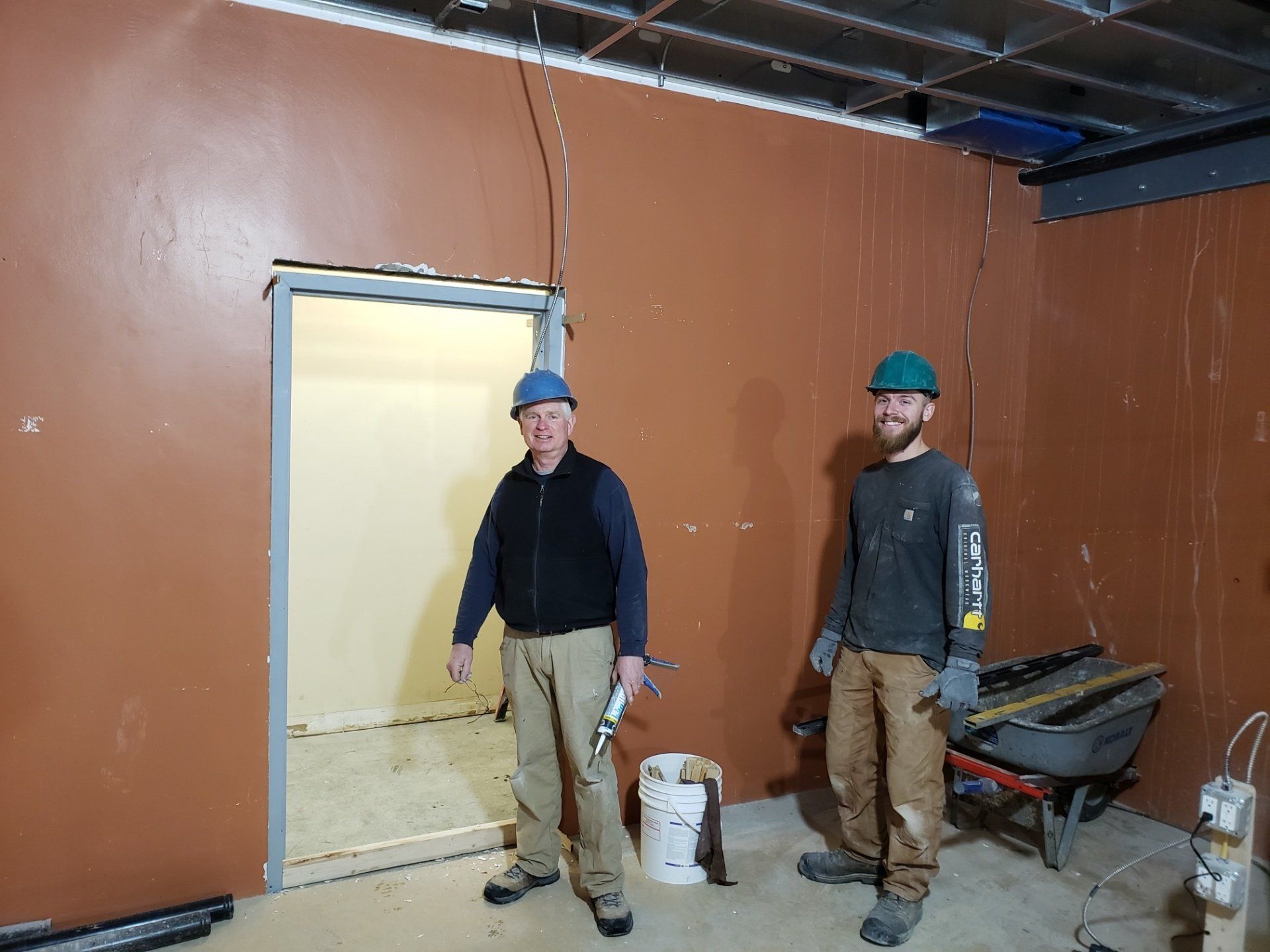 Two workers in hard hats stand inside an unfinished room with brown walls and construction materials.