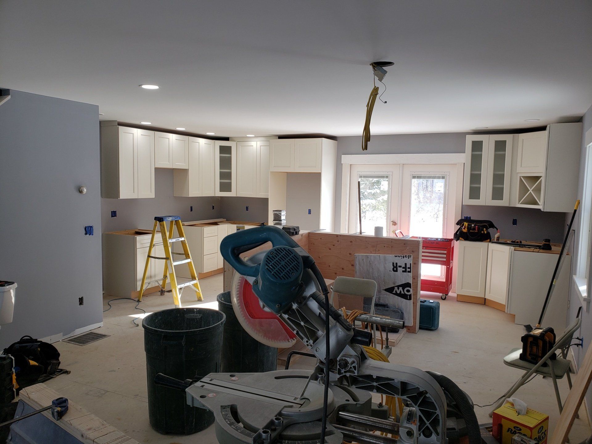 Kitchen renovation in progress with unfinished cabinets, ladders, tools, and a miter saw in the foreground