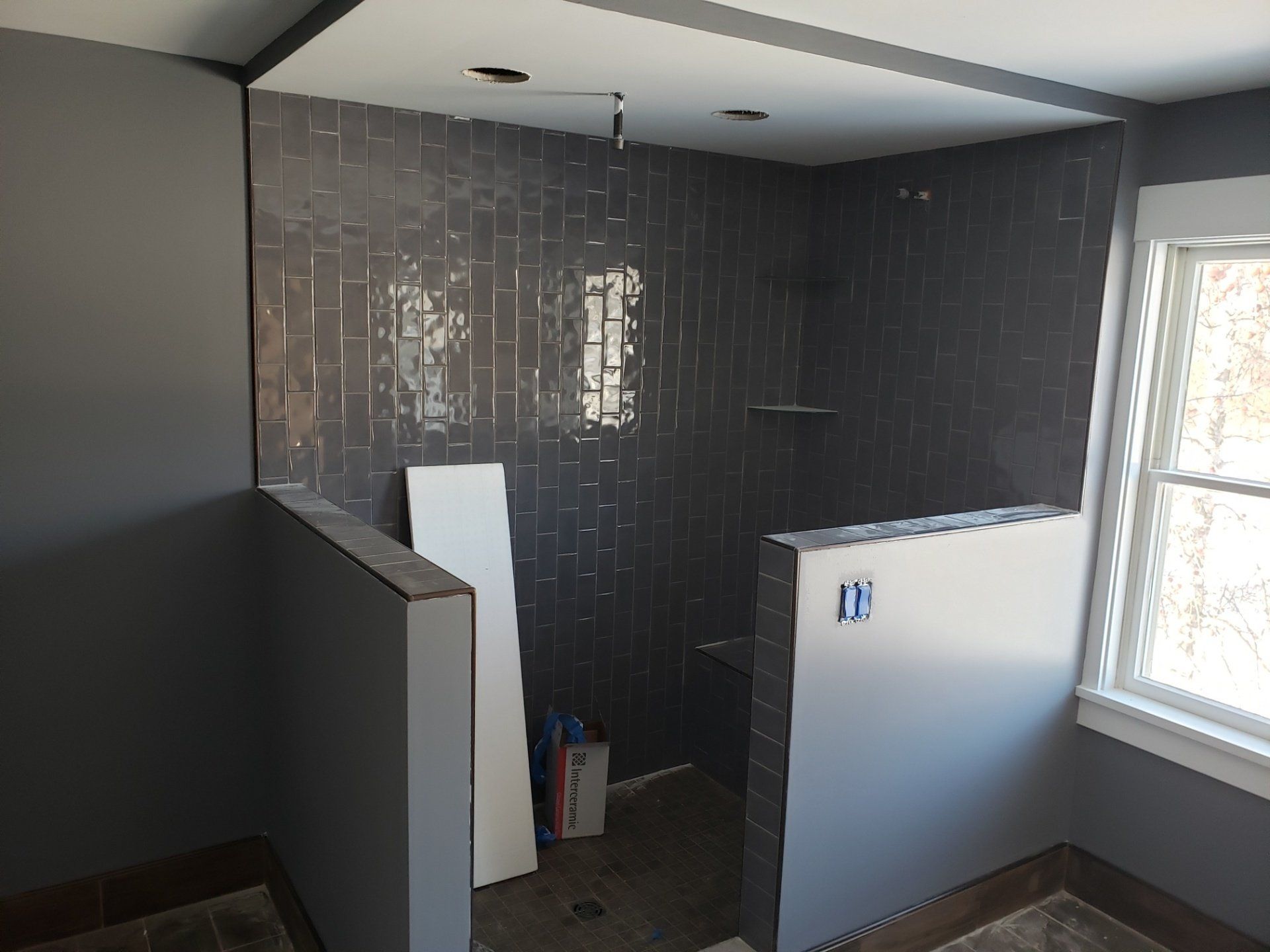 Small unfinished bathroom with dark tile walls, gray partitions, and a window letting in daylight