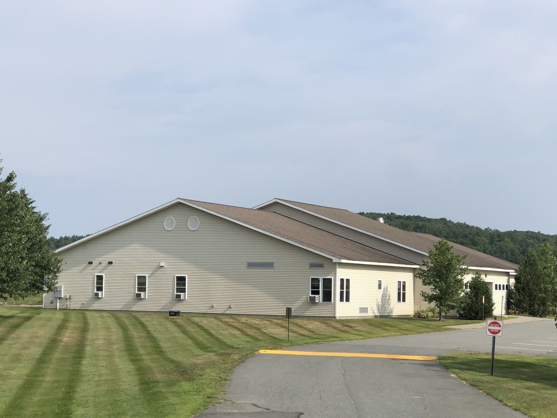 Single-story beige building with a sloped roof beside a mowed lawn and road under a cloudy sky