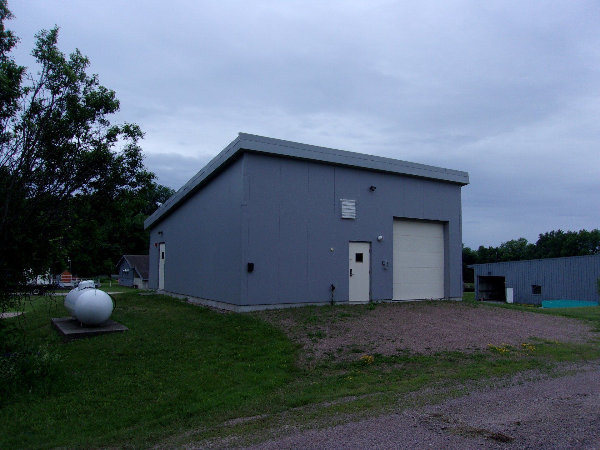 Gray utility building with a roll-up door and propane tank on a gravel lot at dusk