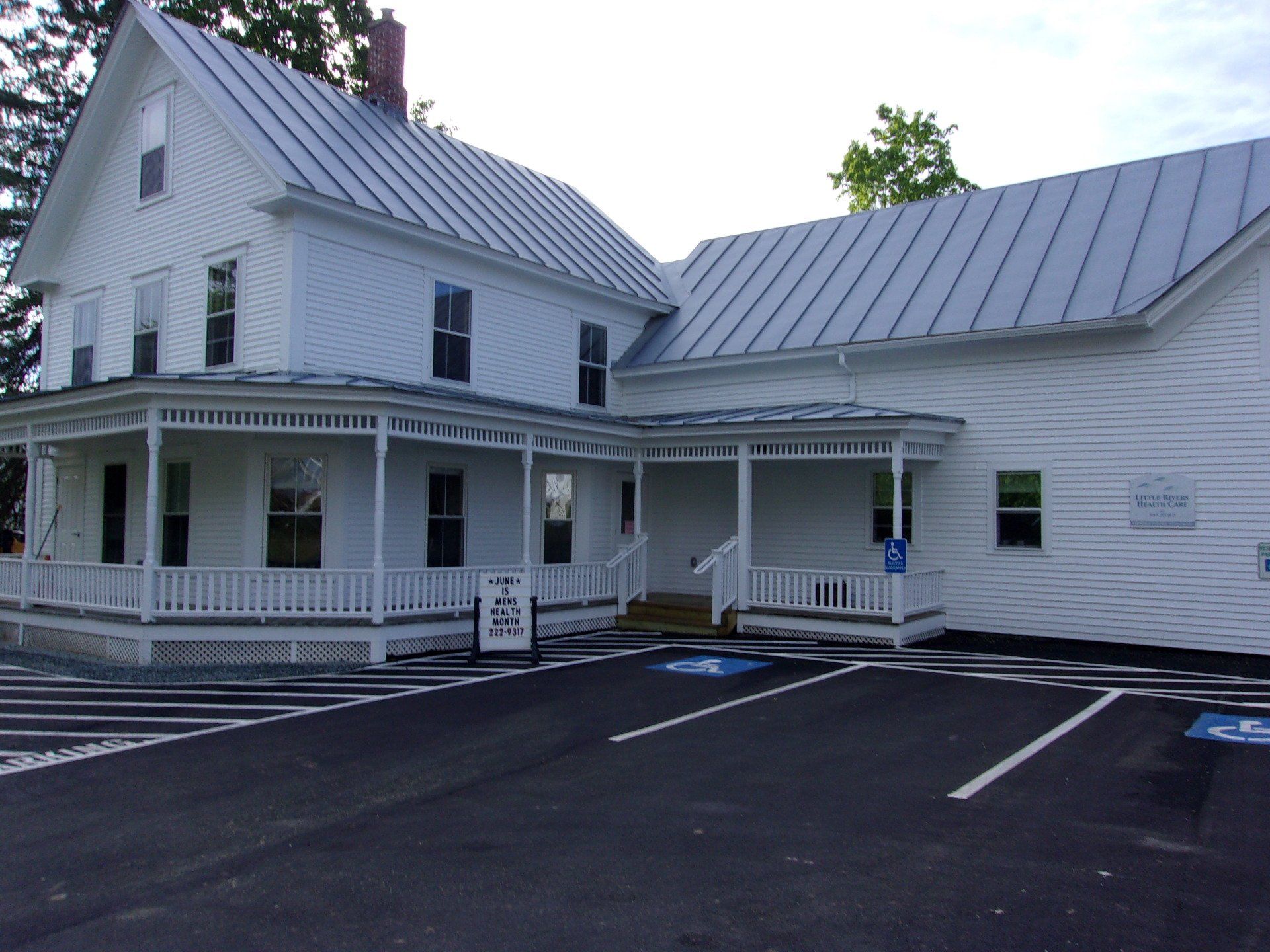White farmhouse-style building with porch and metal roof beside an empty asphalt parking lot