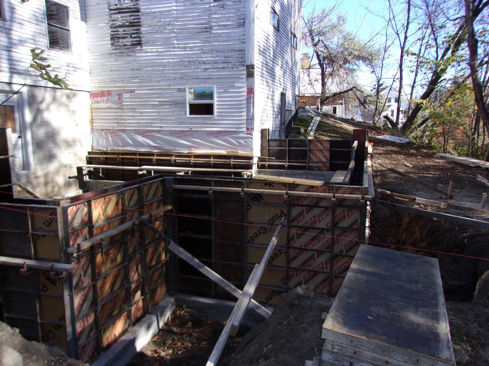 Concrete foundation under construction beside a white house, with wooden forms and scaffolding.