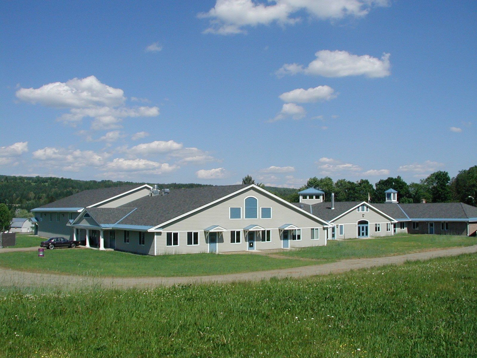 Large white farmhouse with gray roof on a green field under a blue sky with scattered clouds