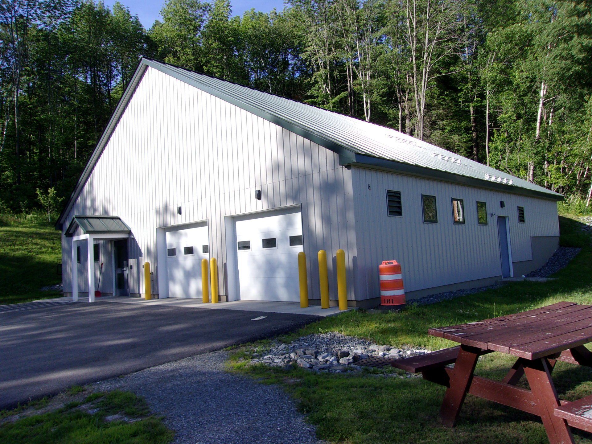 White metal garage building with multiple doors, set beside trees and a picnic table in a sunny area
