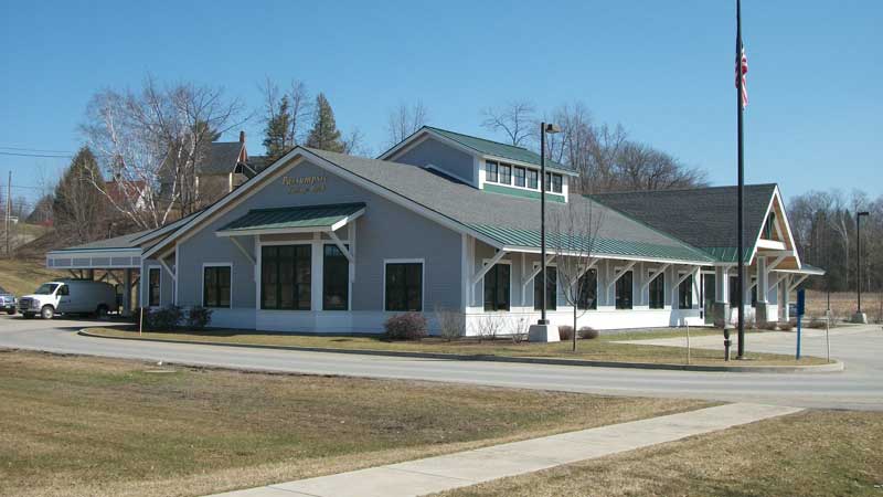 Single-story gray building with green roof and an American flag, seen from a grassy lawn under a clear sky