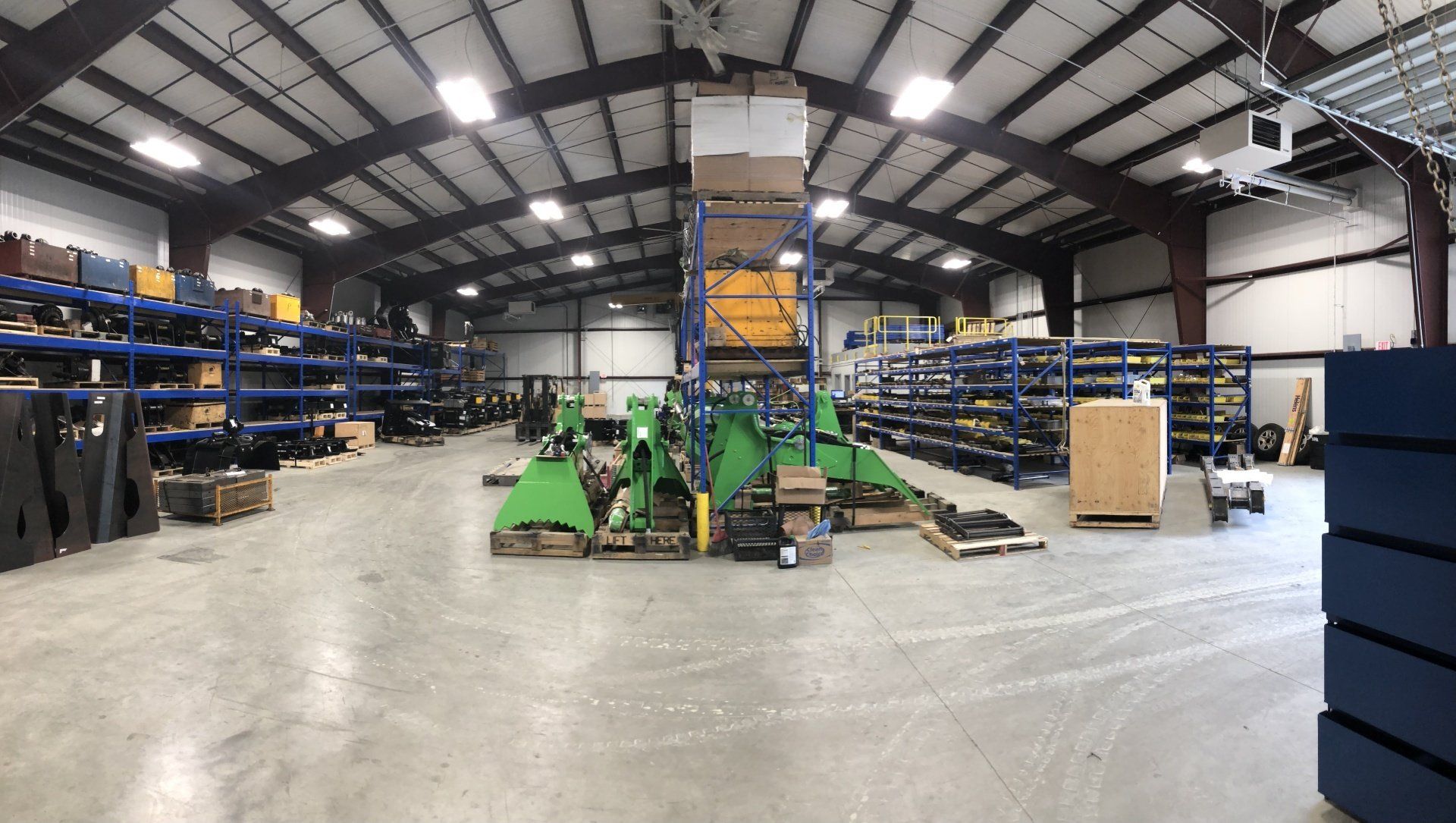Large warehouse interior with blue shelving, a green floor machine, and stacks of boxes under high metal beams